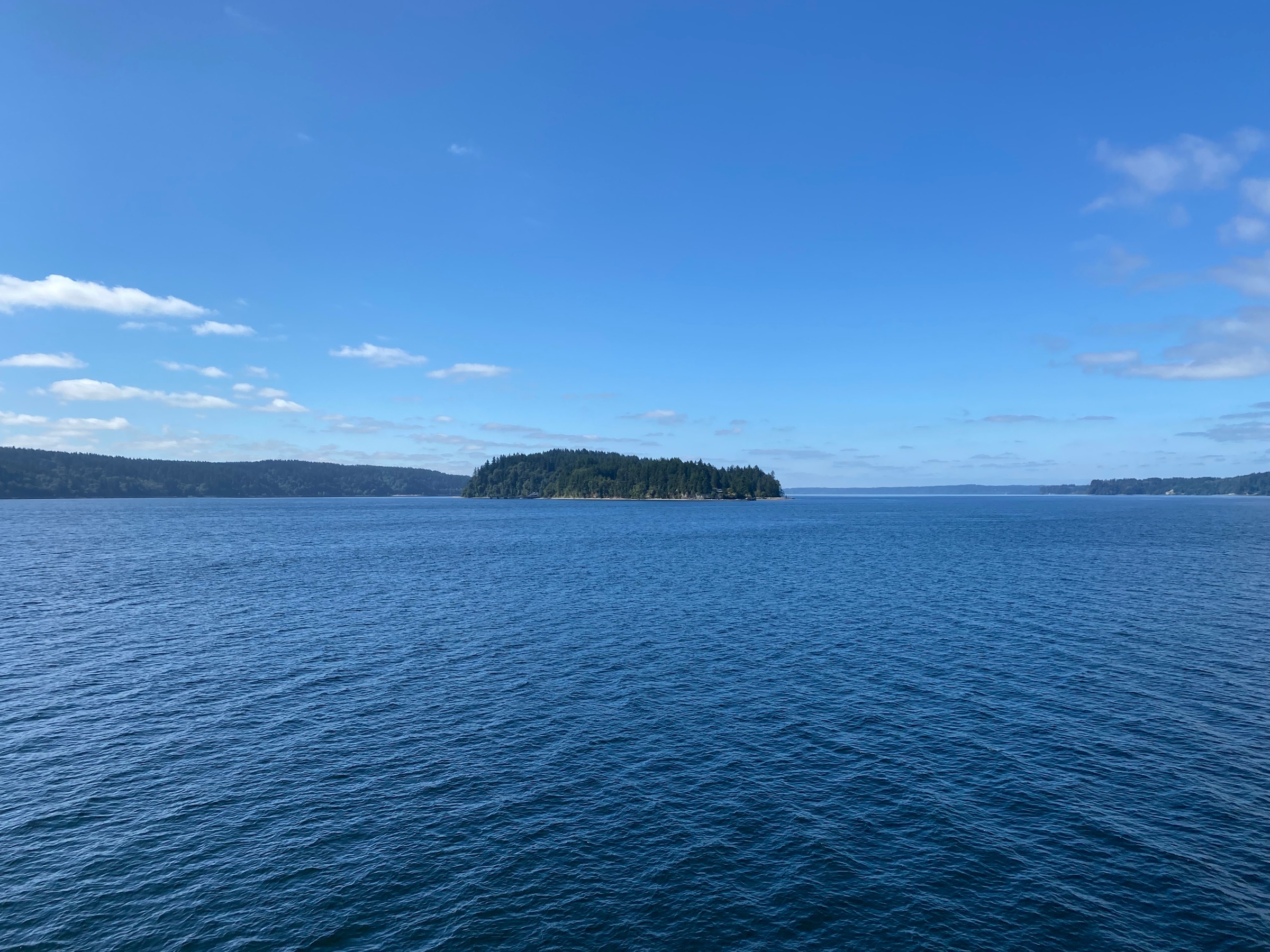Ketron Island seen from the Pierce County ferry on a clear August day, its forested shoreline centered in the distance across deep blue Puget Sound waters.
